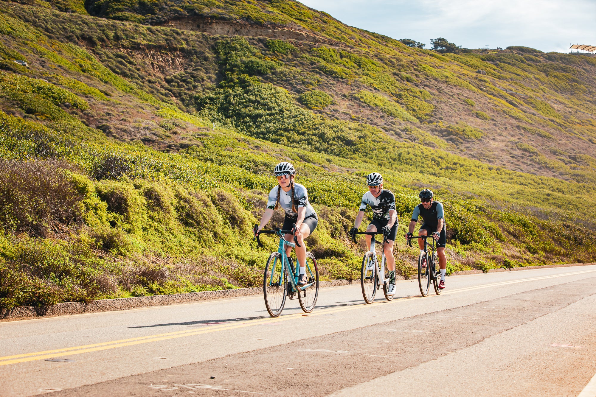 Three cyclists riding down a remote road past a tall grassy hill